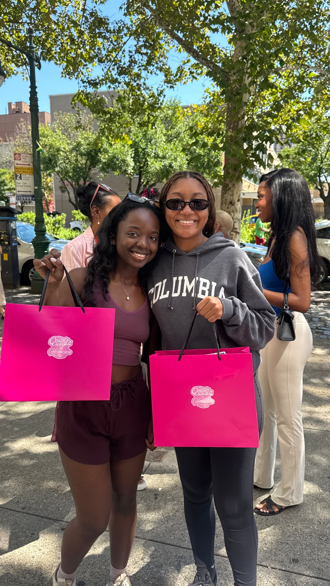 Two young women holding pink Her Voice Project bags on a sunny Harlem sidewalk